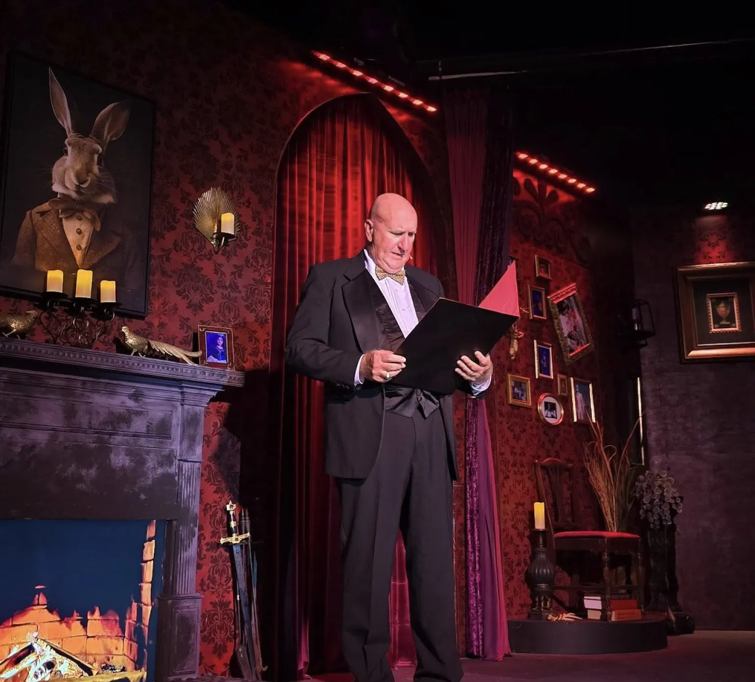 A man in formal attire reads from a folder on a vintage, dimly lit stage with ornate decor and red lighting.