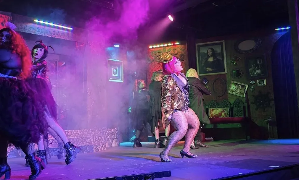 Performers in vibrant costumes dance on a smoky, colorful stage decorated with eclectic framed artwork.