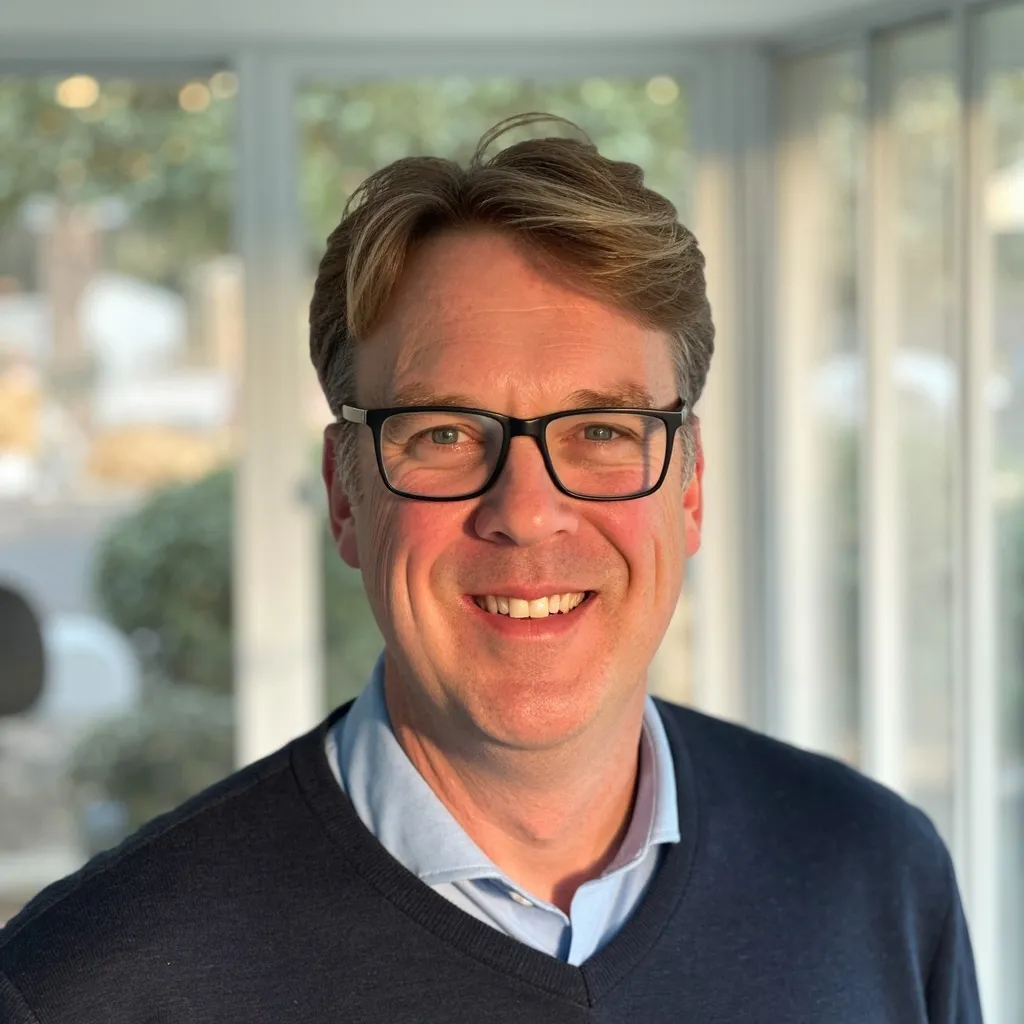 Smiling man with glasses and light brown hair, wearing a blue sweater and collared shirt indoors.