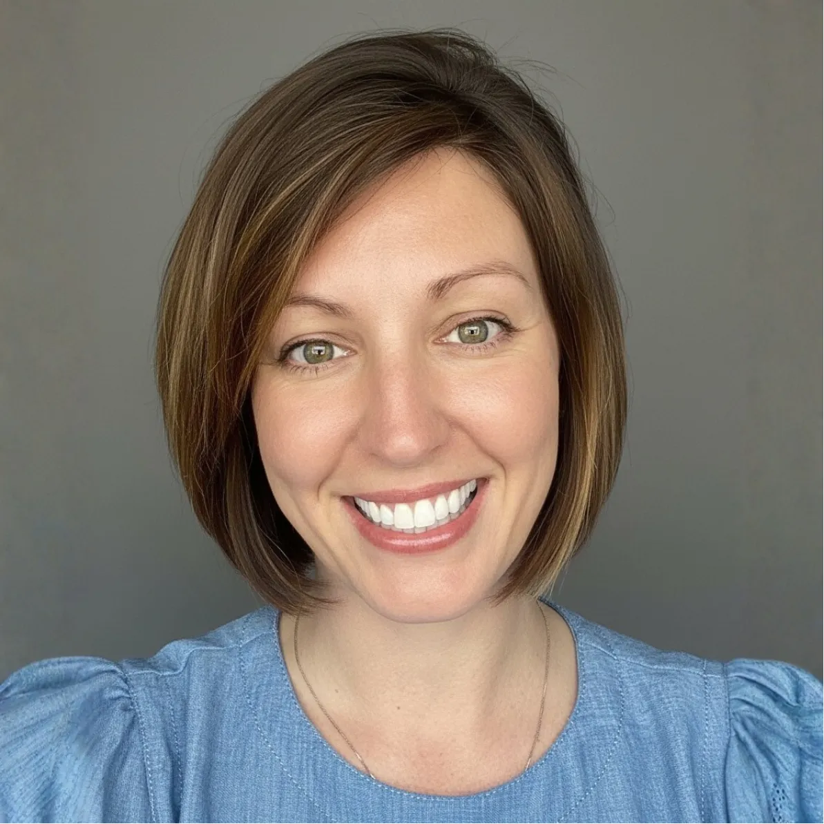 Smiling woman with short brown hair wearing a blue top, posing in front of a plain gray background.