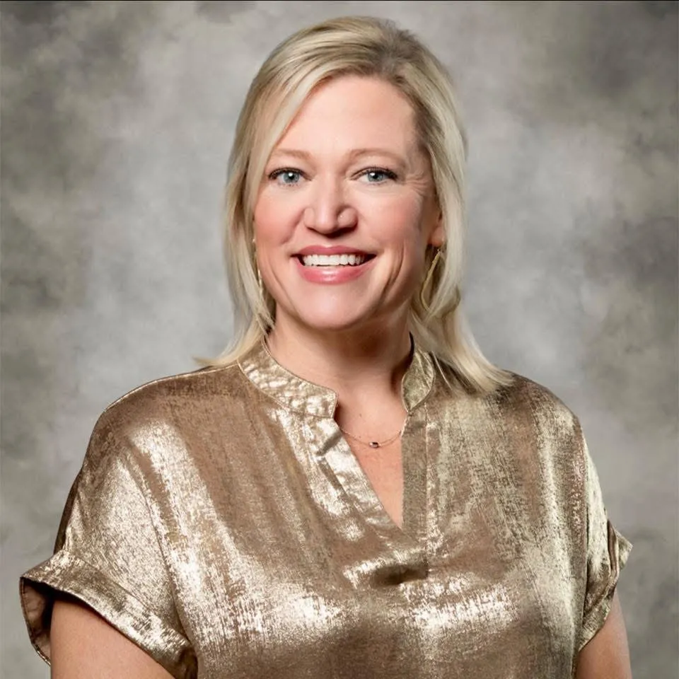 Smiling woman with blonde hair, wearing a shiny gold blouse, standing in front of a gray studio background.