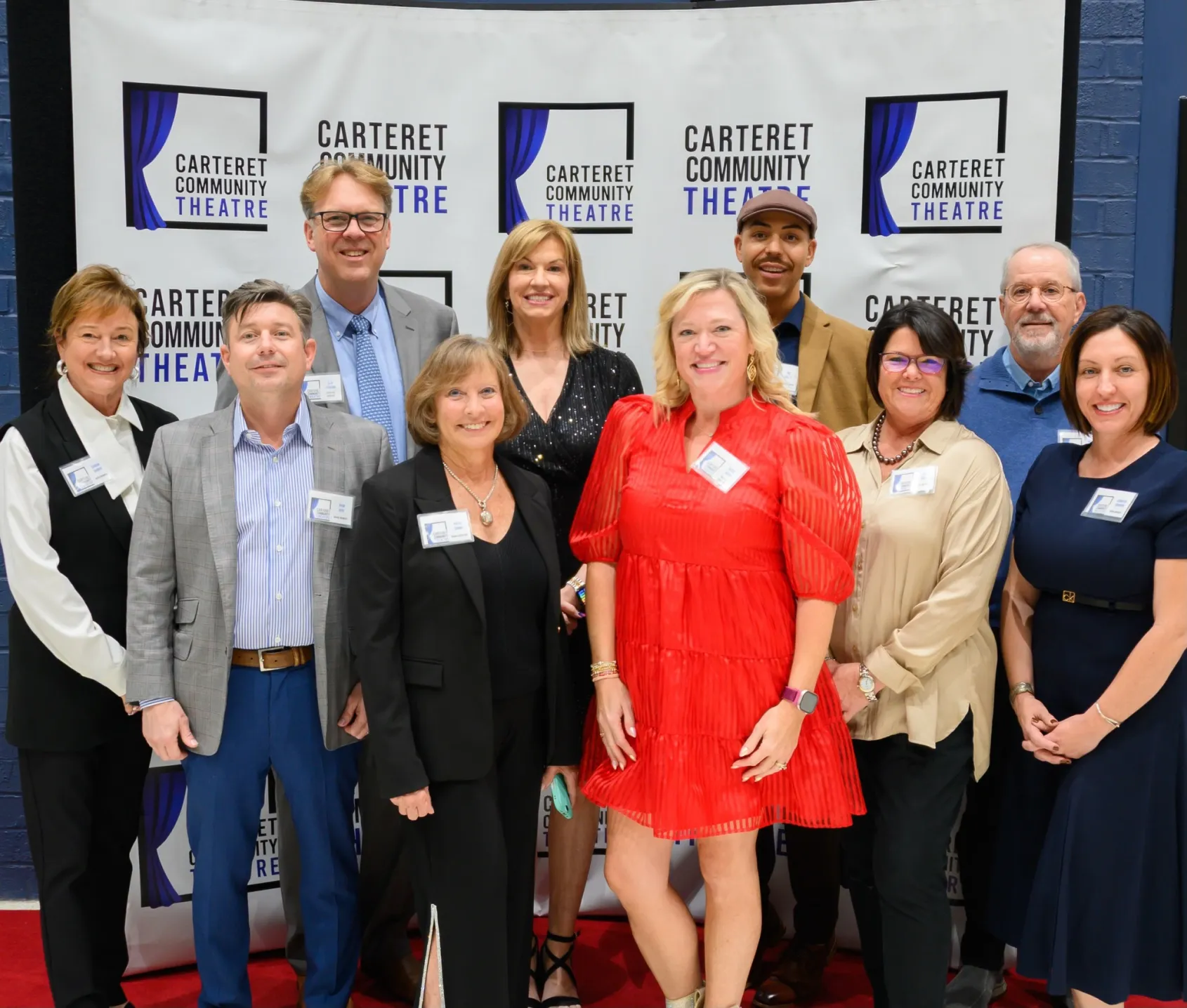 A group of eleven smiling adults poses together in front of a Carteret Community Theatre backdrop.