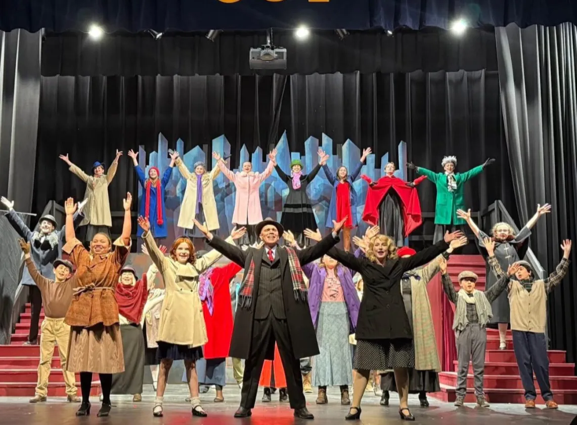 A theater cast in colorful costumes poses on stage with arms raised, in front of a cityscape backdrop.