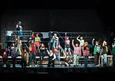 A diverse cast performs on stage, posing energetically in colorful costumes in front of a brick backdrop.