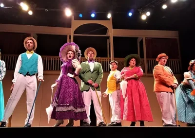 Actors in colorful, vintage costumes perform on stage under bright lights with a simple set in the background.