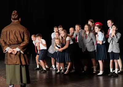 A group of schoolchildren react with surprise to an adult facing them on a stage.