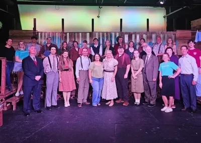 A large group of people in costumes poses on a theater stage with a colorful, rustic set in the background.