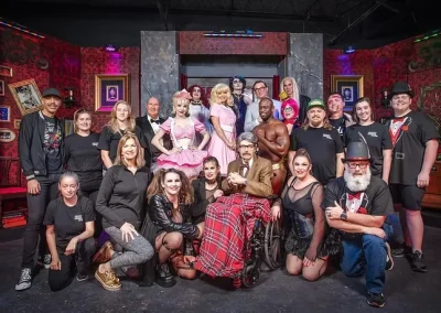 A theater cast and crew pose together in costume on a colorful, decorated stage set.