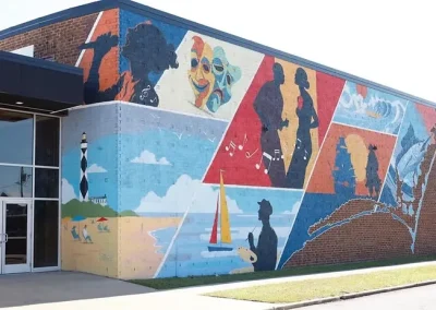 Colorful mural on a brick building depicts people, music, sailing, a lighthouse, and beach scenes.