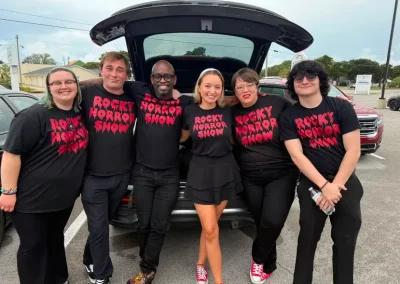Six people stand together smiling in front of a car trunk, all wearing "Rocky Horror Show" shirts.