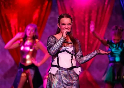 Woman in futuristic costume sings on stage with colorful lights and dancers in metallic outfits behind her.