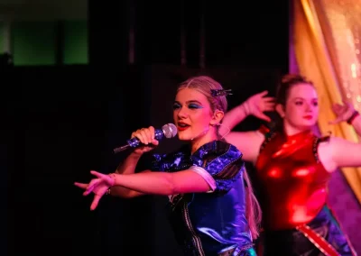 Two performers in vibrant costumes sing and dance on stage with dramatic lighting and gestures.