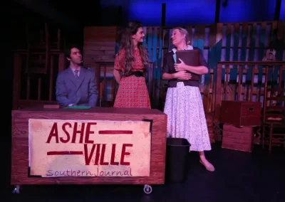 Three actors on stage; two women talking and smiling, one man seated behind a desk labeled "Asheville Southern Journal.
