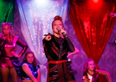 Singer in a colorful stage costume performs with backup dancers under red, green, and white lights.