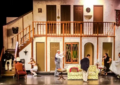 A chaotic theater scene with actors on a set featuring multiple doors, stairs, and a balcony.