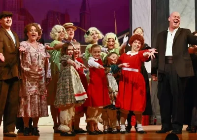 A theater cast in costume, including children in red dresses, sings together on stage with arms outstretched.