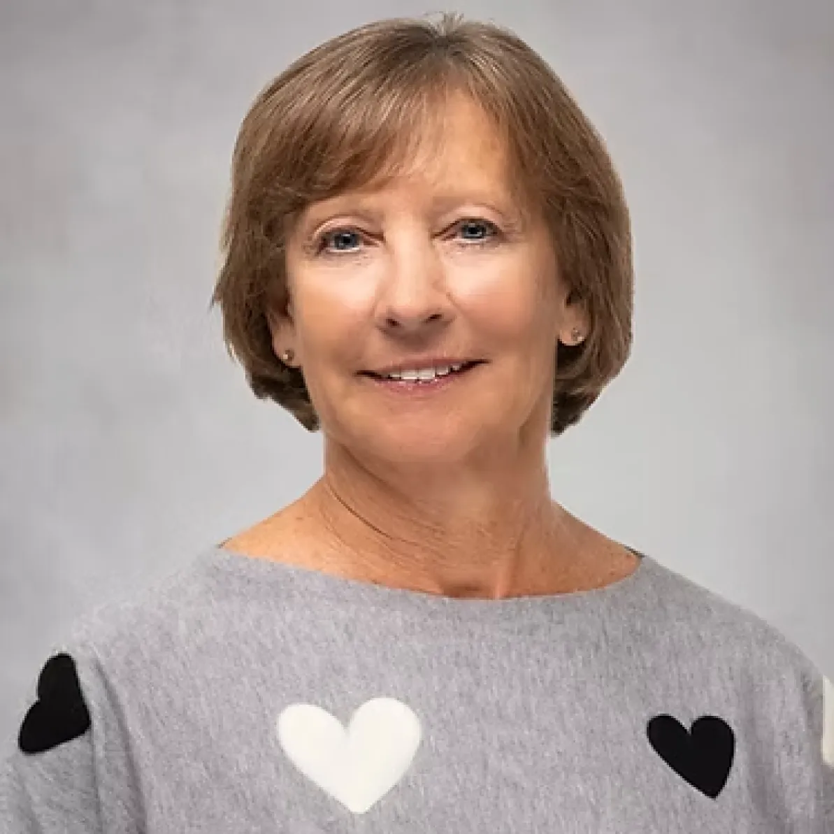 Smiling woman with short light brown hair, wearing a gray sweater with black and white heart shapes.