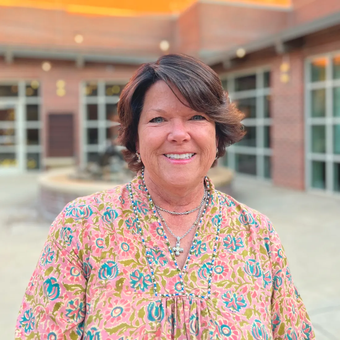 Smiling woman with short brown hair in a floral blouse standing outside a brick building.