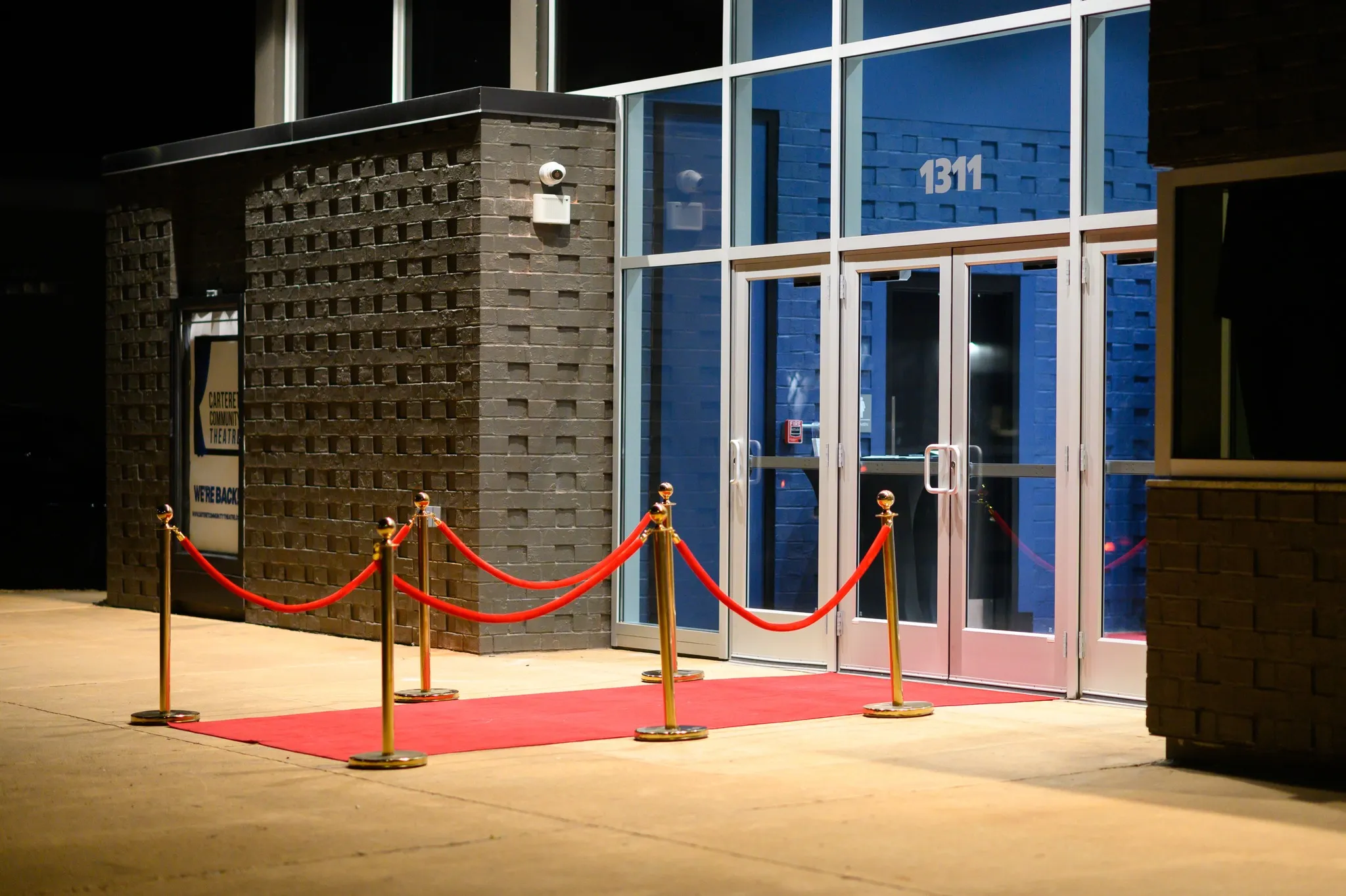 A red carpet with gold stanchions leads to glass doors of a modern brick building at night.