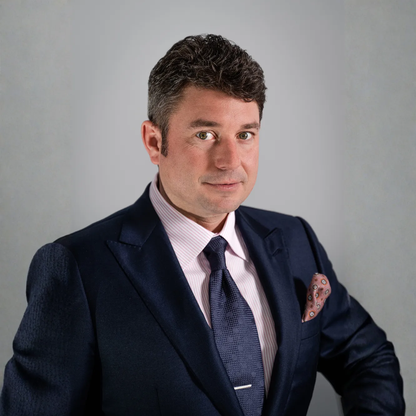 Man in a navy suit, pink shirt, and tie, posing against a plain gray background.