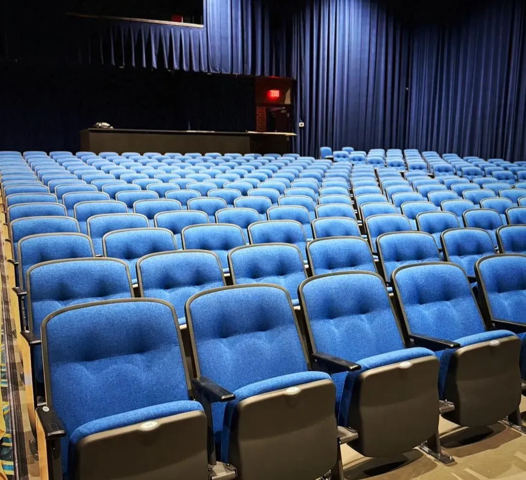 Rows of empty blue theater seats facing a stage with dark curtains and a lit exit sign in the background.