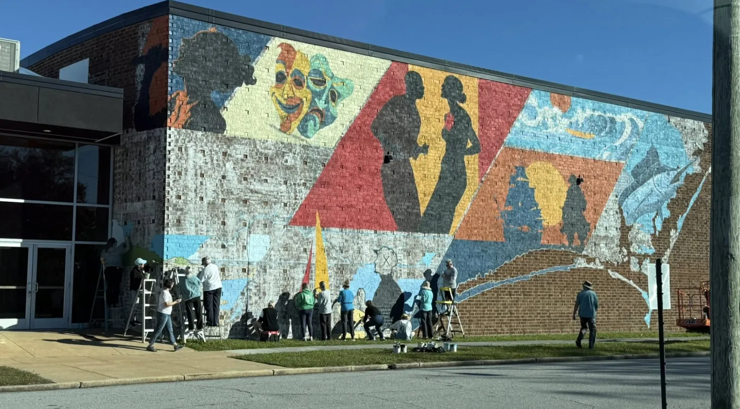 A group paints a colorful mural with silhouettes and faces on the side of a brick building on a sunny day.
