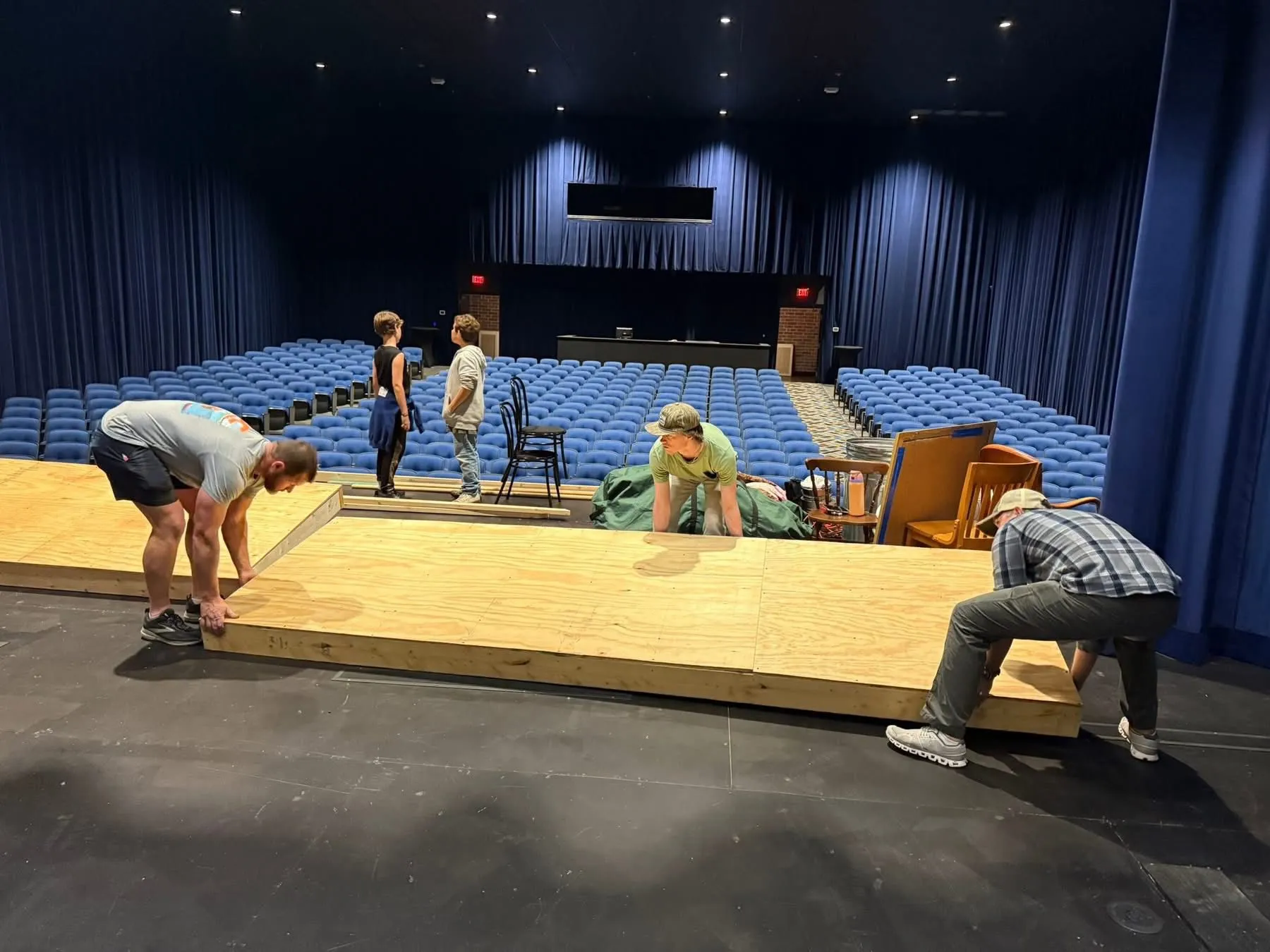 Four people set up a wooden stage platform in a theater with rows of blue seats in the background.