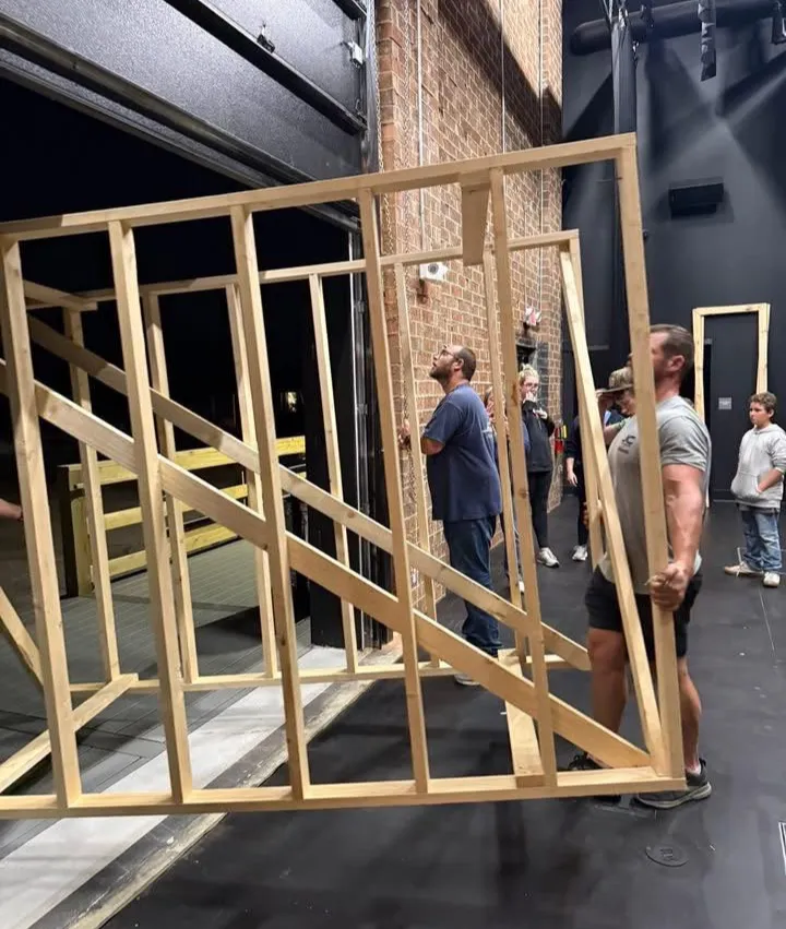 Several people carry a large wooden frame structure inside a building with black floors and brick walls.