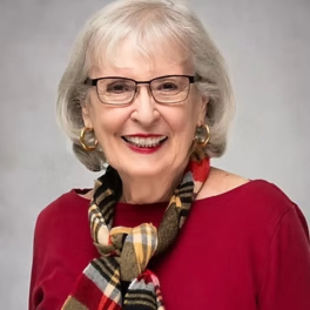 Smiling older woman with gray hair, glasses, red top, and a colorful scarf against a light background.