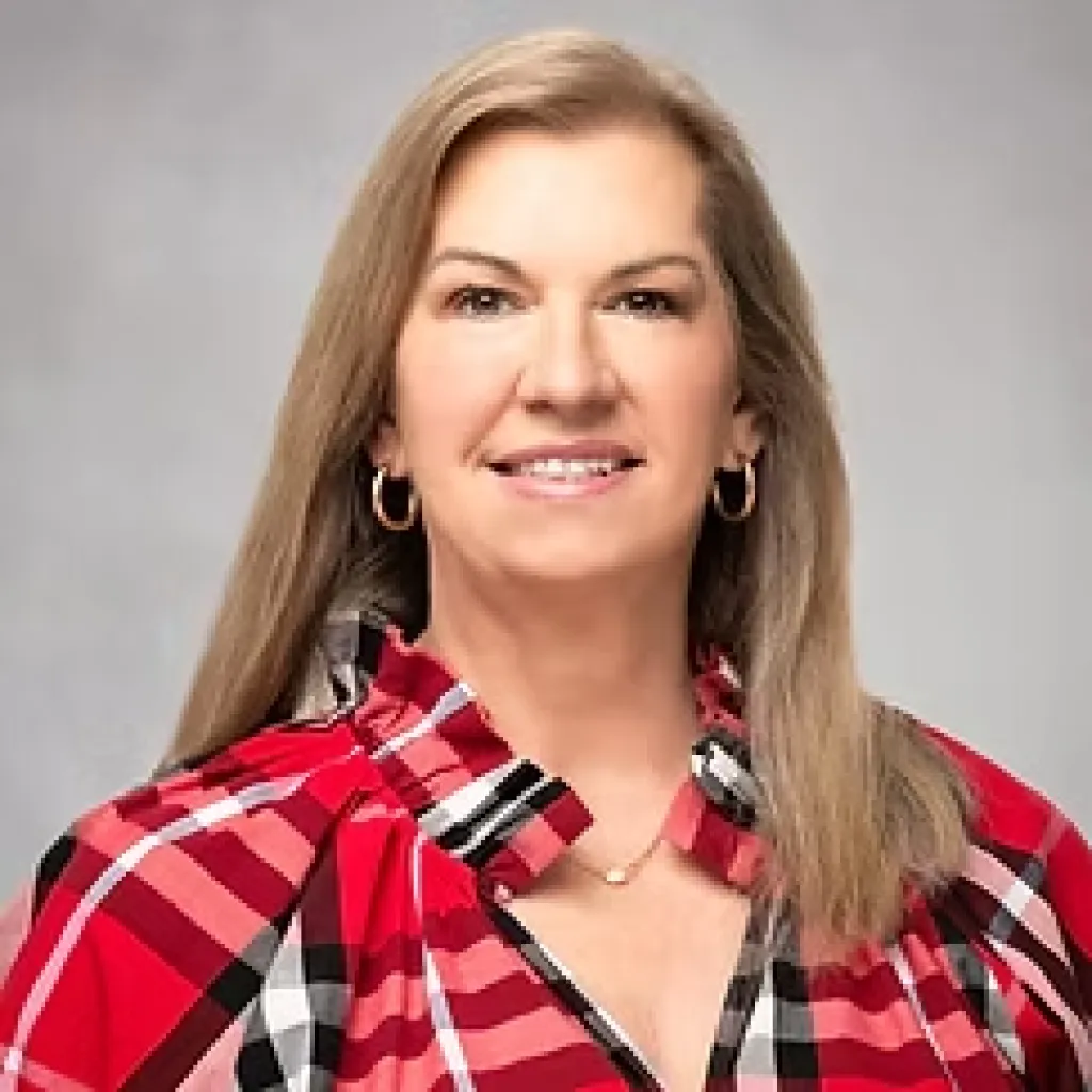 Smiling woman with long hair, wearing a red plaid shirt and gold earrings, posing against a gray background.