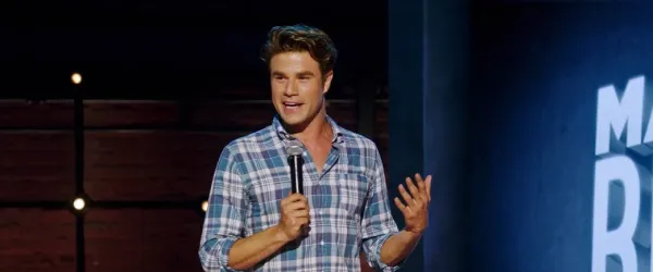 A man in a plaid shirt holds a microphone and speaks on stage during a stand-up comedy performance.