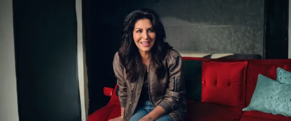 Woman with long dark hair sits on a red couch, smiling, wearing a brown jacket and jeans.