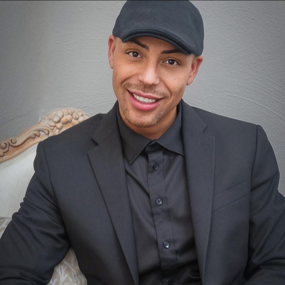 Smiling man in a black suit and cap sits on an ornate chair against a plain gray wall.