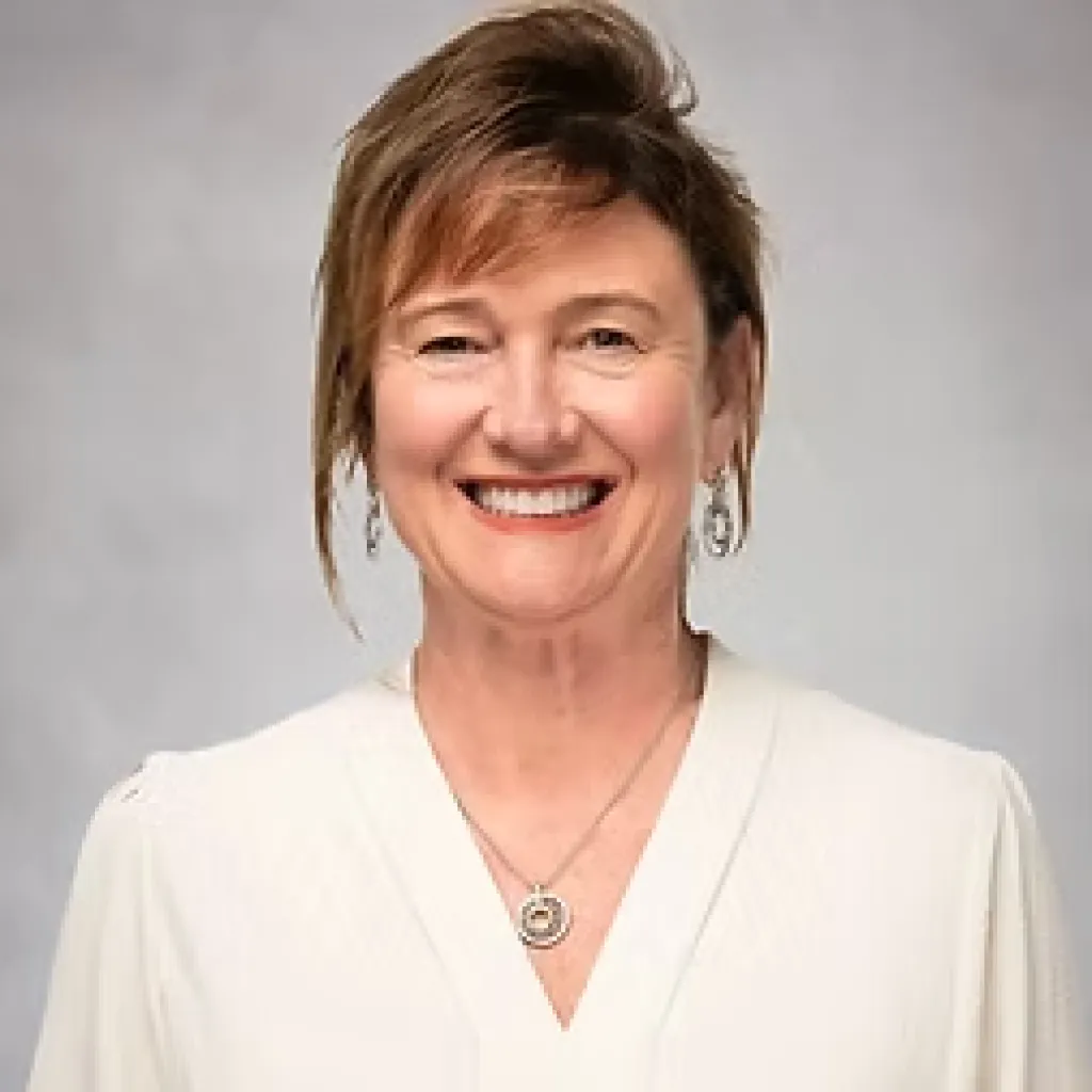 Smiling woman with short brown hair wearing a white top, necklace, and earrings, in front of a gray background.