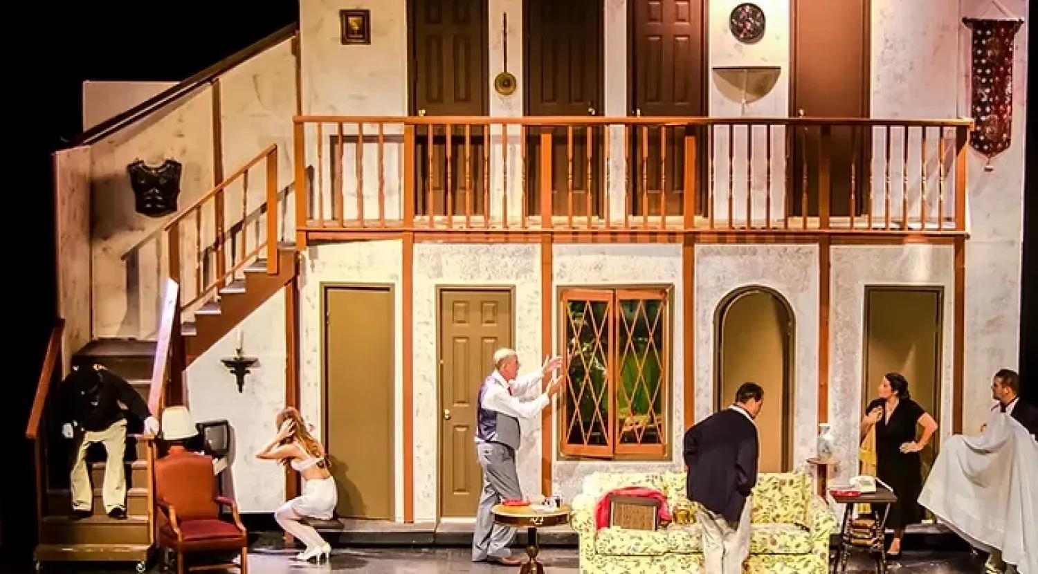 Actors perform a comedic scene in a colorful living room set with multiple doors and a staircase.