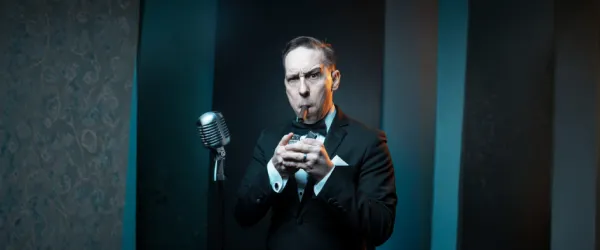Man in tuxedo drinking from a glass with a straw, standing beside a vintage microphone on stage.