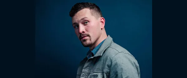 A man with short hair and a goatee looks at the camera, wearing a gray jacket against a blue background.
